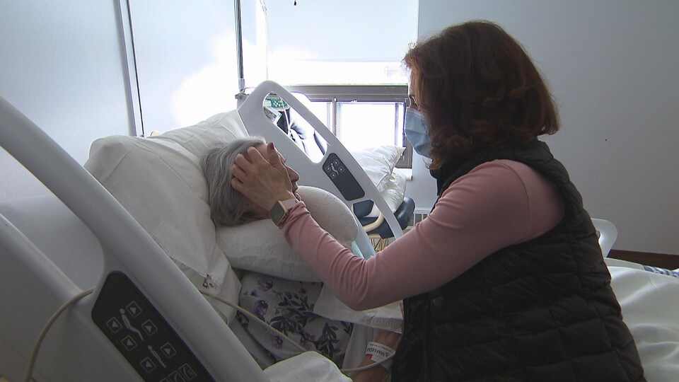 Marcelle dans un lit médicalisé, à côté de sa fille qui lui caresse les cheveux.