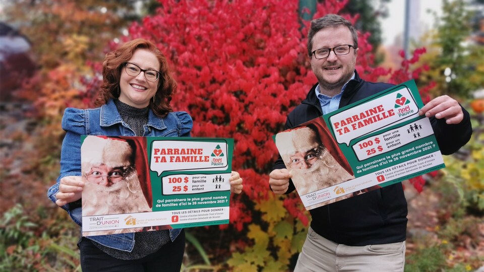 Isabelle Bombardier et Mathieu Gélinas tiennent une affiche de la campagne « Parraine ta famille » devant un buisson.