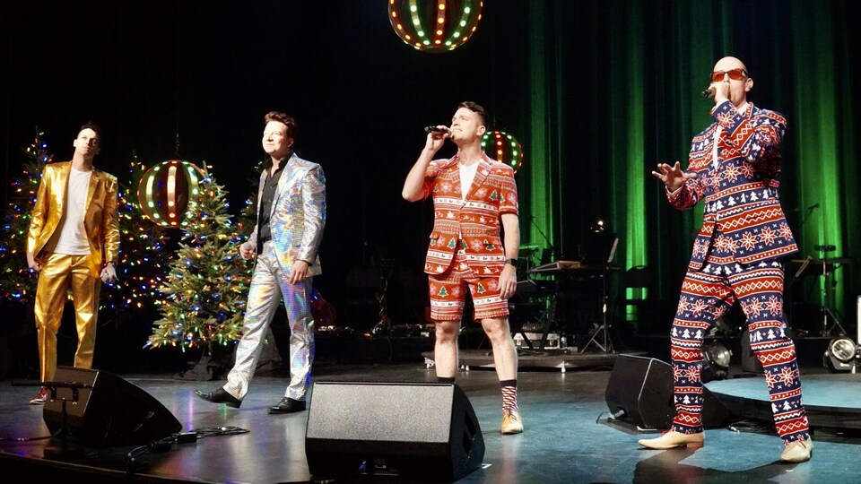 Quatre hommes aux complets colorés chantent sur une scène.