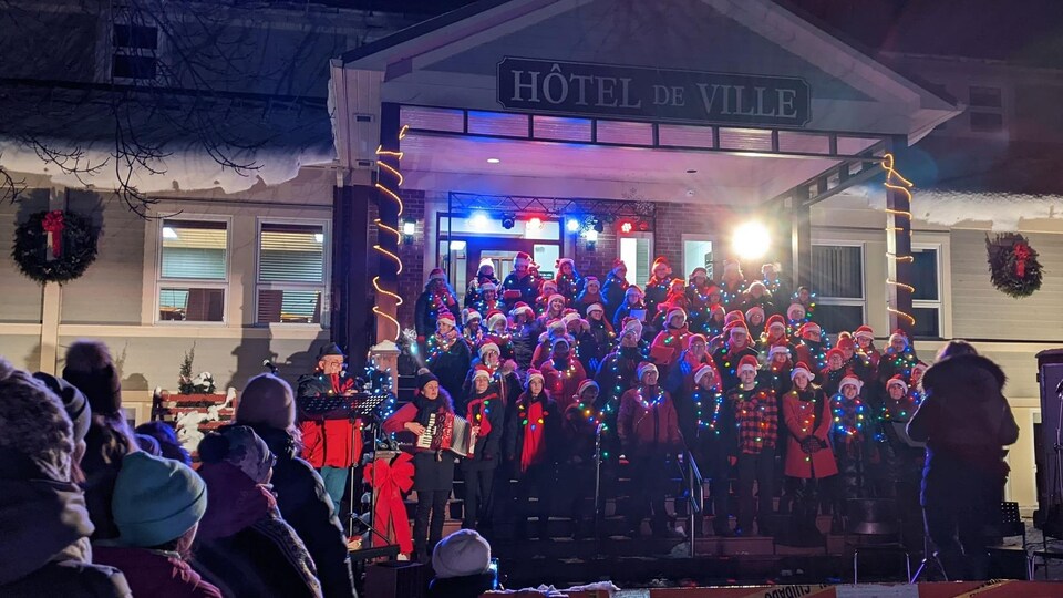 Une chorale sur les marches de l'Hôtel de Ville de New Richmond. Les chanteurs portent des tuques de Noël et des lumières de Noël.