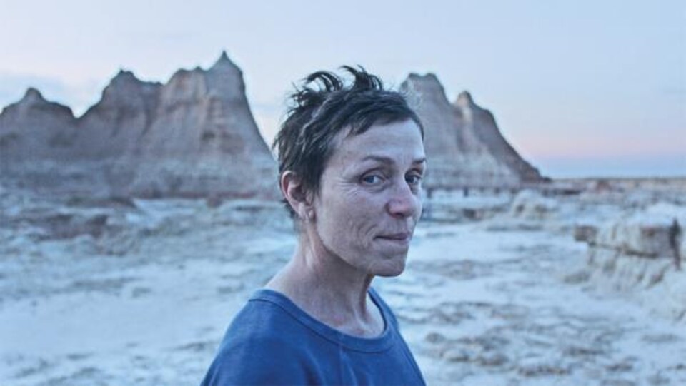 Une femme regarde vers l'objectif dans un désert, avec des montagnes en arrière-plan.