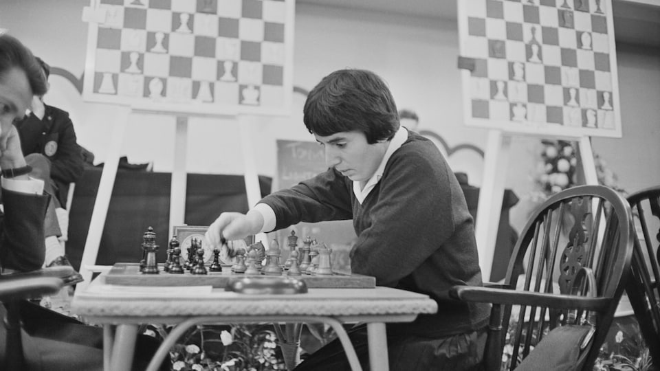 Dans une photo en noir et blanc, une femme est assise devant un jeu d'échecs face à un adversaire et elle s'apprête à déplacer une pièce sur l'échiquier.