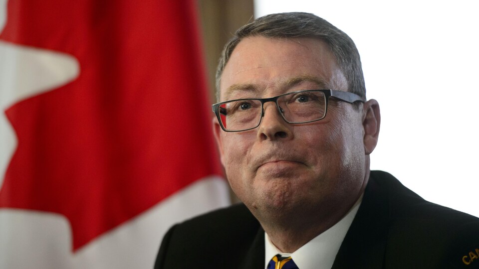 Mark Norman sourit, la bouche fermée, alors qu'on voit un drapeau canadien derrière lui.