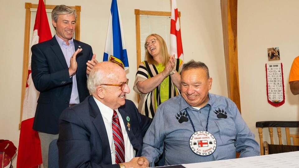 Norman Sylliboy et Arthur LeBlanc sont assis et se serrent la main. Tim Houston et Karla MacFarlane sont assis derrière et applaudissent.