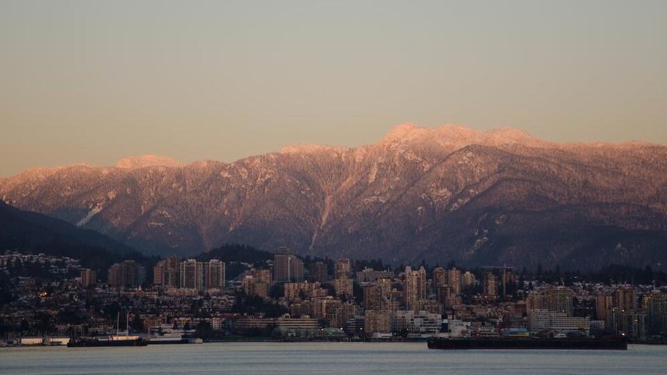 D'imposantes montagnes enneigées avec un lever de soleil et une ville avec des gratte-ciel devant une étendue d'eau.