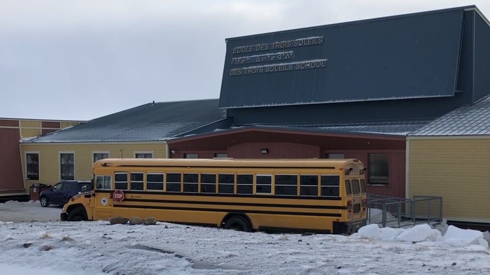 Un autobus scolaire est stationné devant un édifice architectural dont le nom est affiché en trois langues.