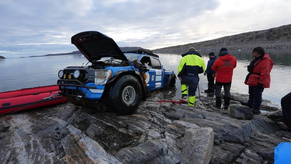 Un groupe de quatre personnes se trouvent à côté d'un camion au capot ouvert et à moitié dans l'eau, moitié sur la rive, en août 2022 au Nunavut.