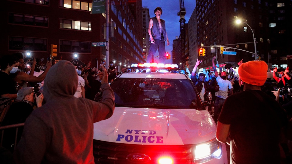 Un manifestant debout sur une voiture de police entourée de nombreux manifestants dans une rue.