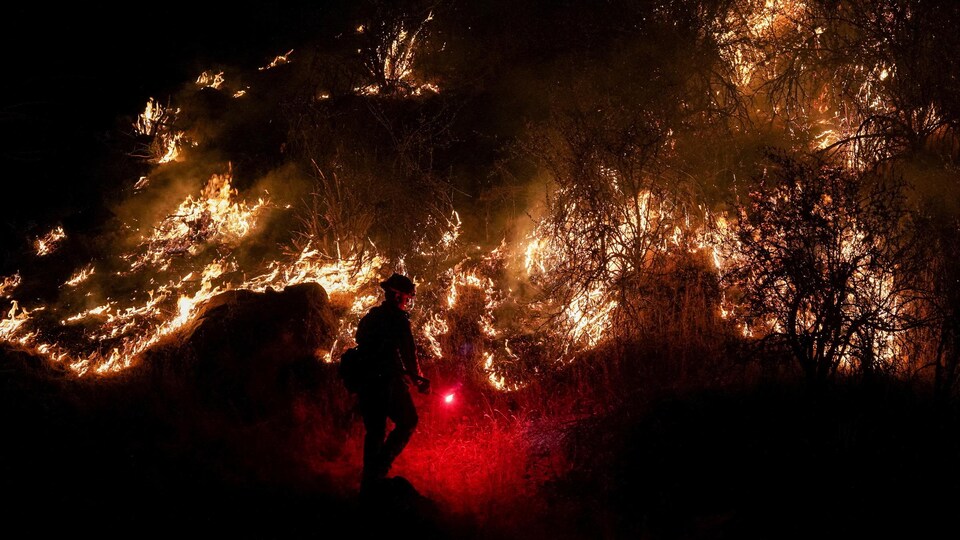 Un pompier allume un feu de brûlage dirigé alors qu'un incendie agricole brûle devant lui la nuit.