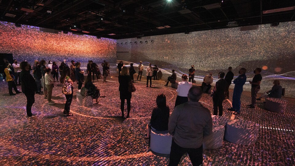 Des personnes sont immobiles au sein d'une grande pièce où sont projetés des milliers de petits faisceaux lumineux sur les murs et le plancher.