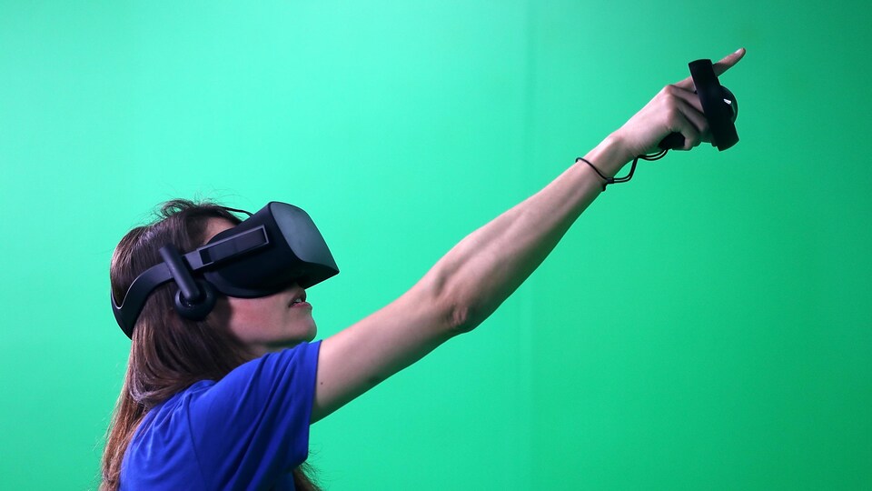 Une femme portant un casque de réalité virtuelle pointe le ciel, devant un écran vert. 