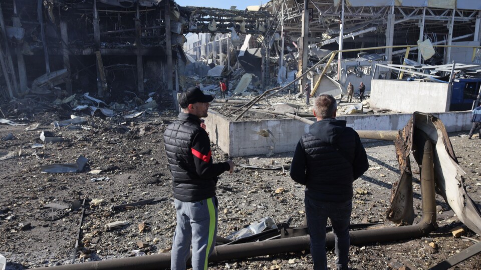 Deux hommes, de dos, observent un immeuble complètement détruit. 