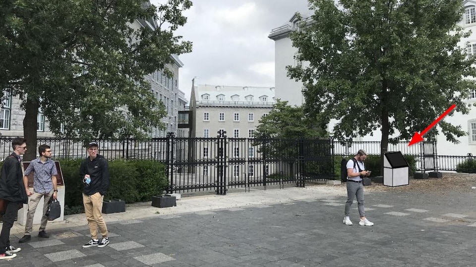 Une stèle est pointée par une flèche rouge dans la cour du Séminaire de Québec, dans une scène d'été, alors que des étudiants déambulent tranquillement dans le secteur