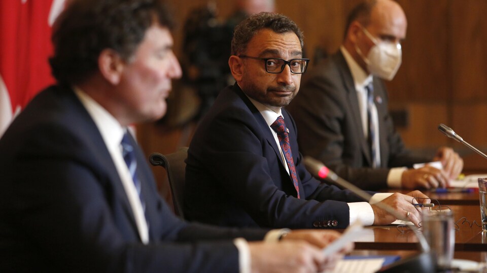 Les trois ministres sont assis à une table, derrière des micros. Omar Alghabra fixe la caméra.