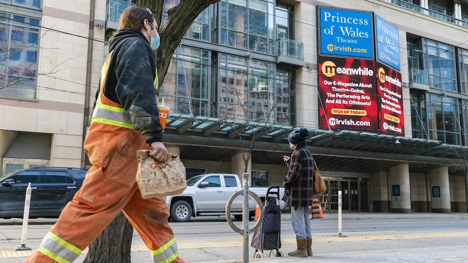 Une femme attend le tramway devant le théâtre Princess of Wales, au centre-ville de Toronto. Un travailleur de la construction marche sur le trottoir.