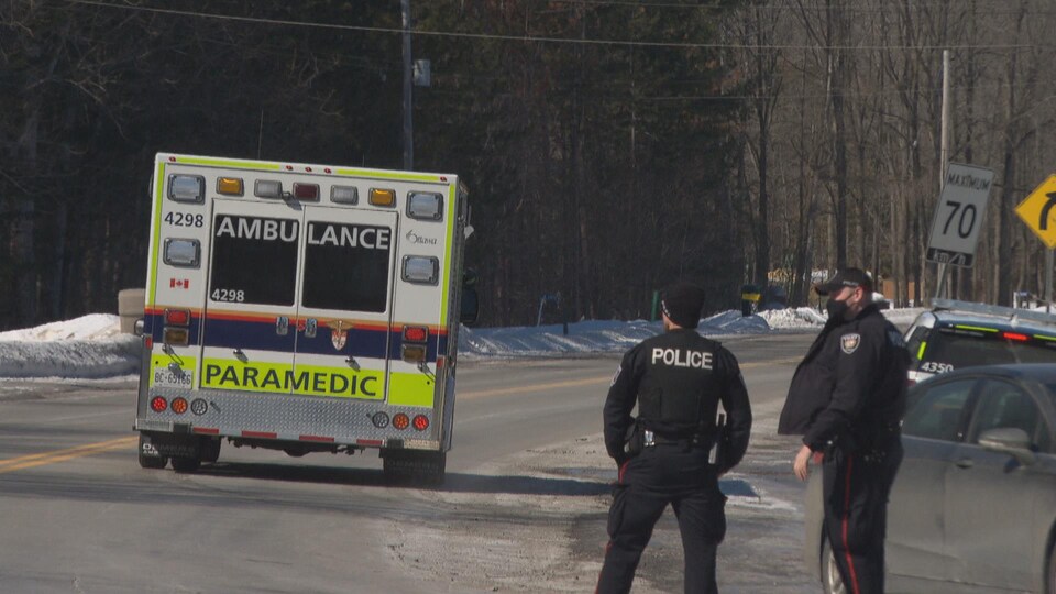 Deux policiers se tiennent l'un à côté de l'autre en bord de route pendant qu'une ambulance s'éloigne.
