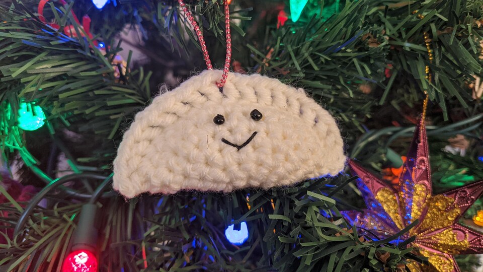 Dans un sapin de Noël tout illuminé, gros plan sur un ornement en laine représentant un pierogi avec deux yeux et une bouche qui sourit.