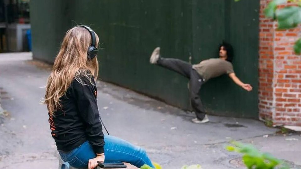 Une femme assise avec des écouteurs regarde un homme effectuer un mouvement de danse dans une ruelle.
