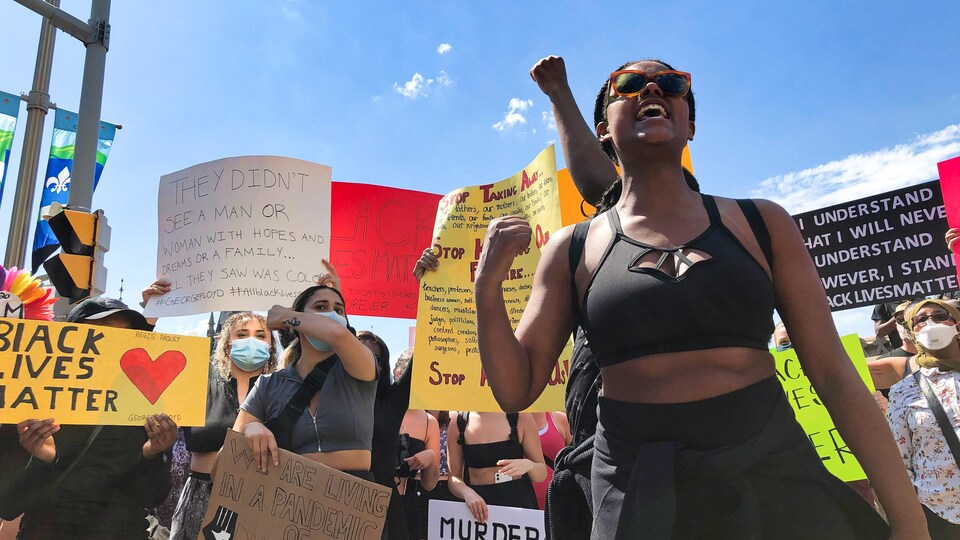 Des dizaines de manifestants semblent crier, plusieurs tenant des pancartes sur lesquelles sont écrits divers messages antiracistes en anglais. 