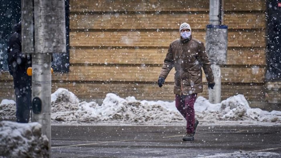 Un piéton masqué traverse une rue lors pendant une averse de neige.