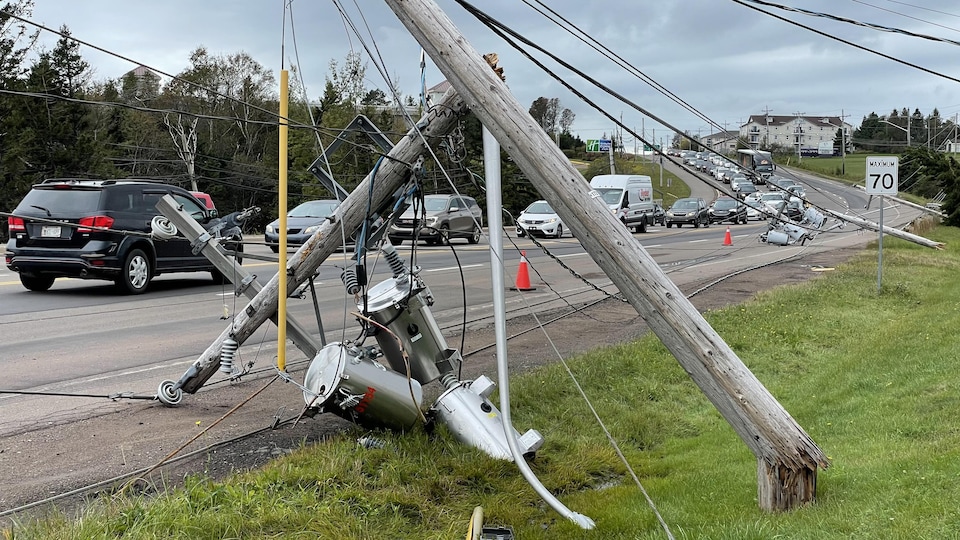 Des poteaux électriques brisés et des lignes à haute tension abattues au bord d'une route sur laquelle des automobiles circulent.