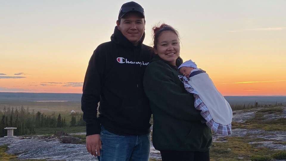 Au soleil couchant à l'extérieur, Paasa Lemire tient son bébé dans ses bras, au côtés de son conjoint.