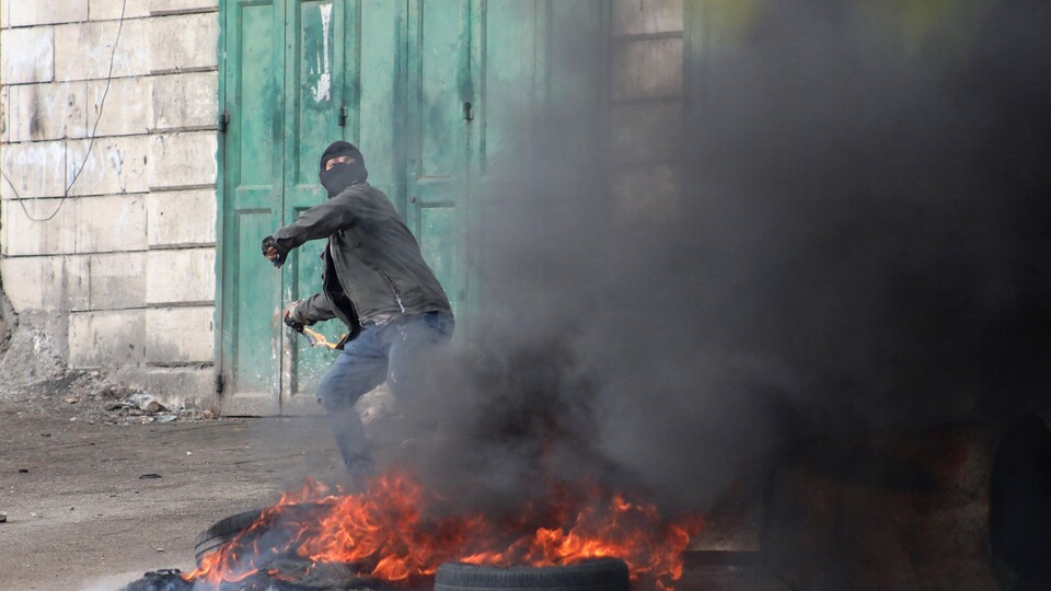 Un manifestant cagoulé lance une pierre avec une fronde derrière un pneu en flammes.