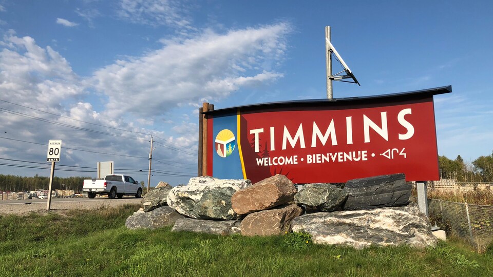 Une camionnette blanche passe près d'une pancarte qui souhaite la bienvenue à Timmins en trois langues.