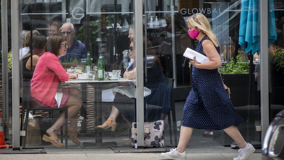 Une personne portant un masque passe devant la terrasse d'un restaurant où sont attablés des gens.