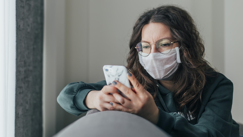 Une femme portant un masque regarde l'écran de son téléphone portable.