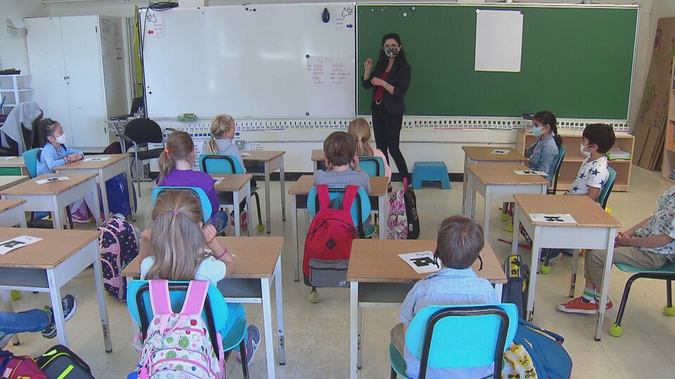 Une femme avec un masque parle aux élèves dans une salle de classe. Certains enfants portent un masque.