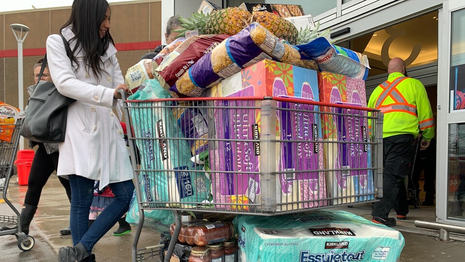 Une femme avec son panier d'épicerie rempli de provisions : mouchoirs, sauces, pâtes, fruits et essuie-tout à la sortie de l'épicerie.