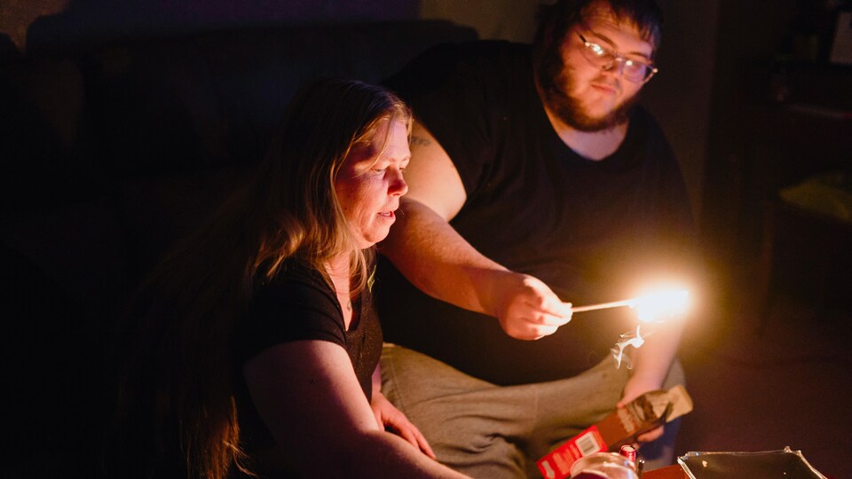 Un homme et une femme sont assis à l'intérieur dans le noir devant des bougies pendant une panne d'électricité. L'homme tient une longue allumette et s'apprête à allumer une autre chandelle.