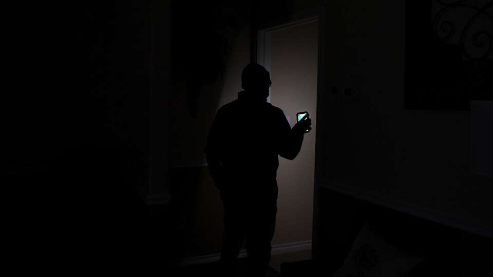Un homme dans un domicile marche dans la quasi-obscurité en s'éclairant avec la lumière de son téléphone pendant une panne d'électricité.