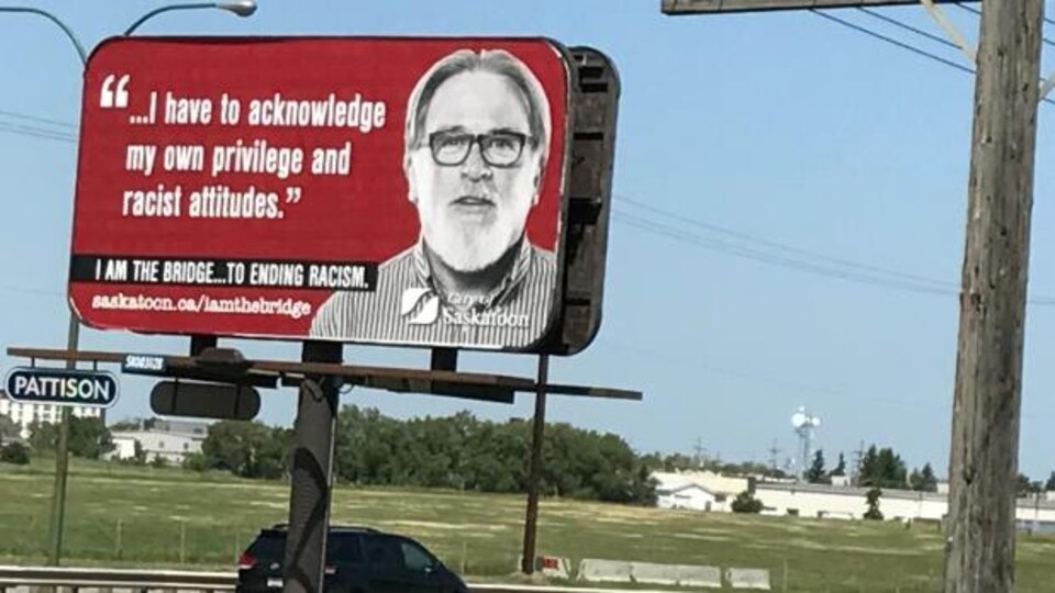 Un panneau publicitaire en rouge, avec un homme portant des lunettes, dit en anglais qu'il reconnait ses propres privilèges et ses attitudes racistes.