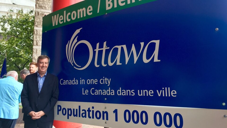 Jim Watson devant un panneau d'accueil de la ville d'Ottawa où la population de 1 million est indiquée et où se trouve le slogan «le Canada dans une ville».