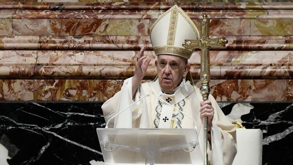 Coiffé d'une mitre et vêtu d'une soutane, le pape tient une grosse croix dans sa main gauche. Il prononce un discours.