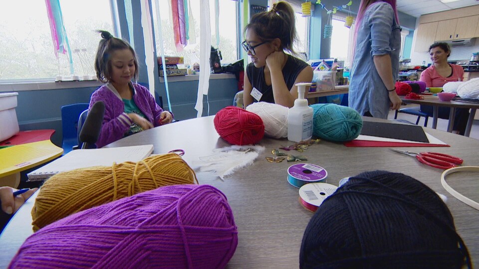 Une fillette et une jeune femme sont assises à une table, sur laquelle sont posées des pelotes de laine, des ciseaux et d'autres accessoires.