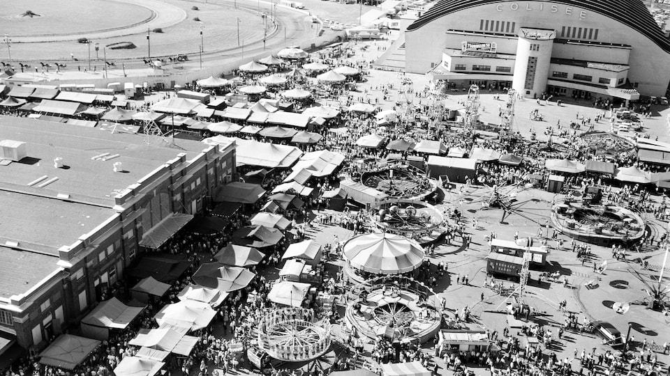 Tentes, manèges et foule dense durant une belle journée d'été, en pleine exposition sur la grande place du Colisée durant les années 1950.