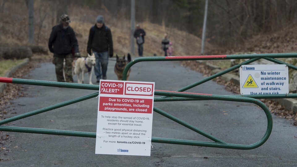 Des gens font marcher leurs chiens dans un sentier à High Park, derrière une enseigne qui précise que certaines installations sont fermées.