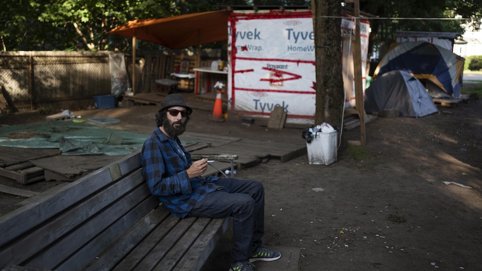 Un homme avec une chemise à carreaux bleue est assis sur un banc de parc. Derrière lui, on voit les abris en bois et les tentes de personnes sans-abri qui y dorment.
