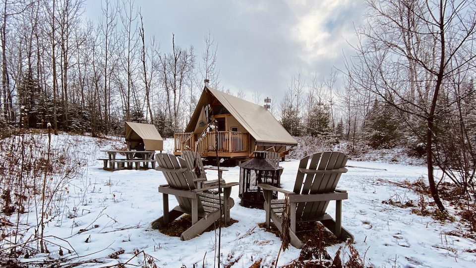 Des chaises Adirondack autour d'un foyer extérieur sont en avant-plan. On voit au loin une petite habitation qui s'apparente à un chalet.