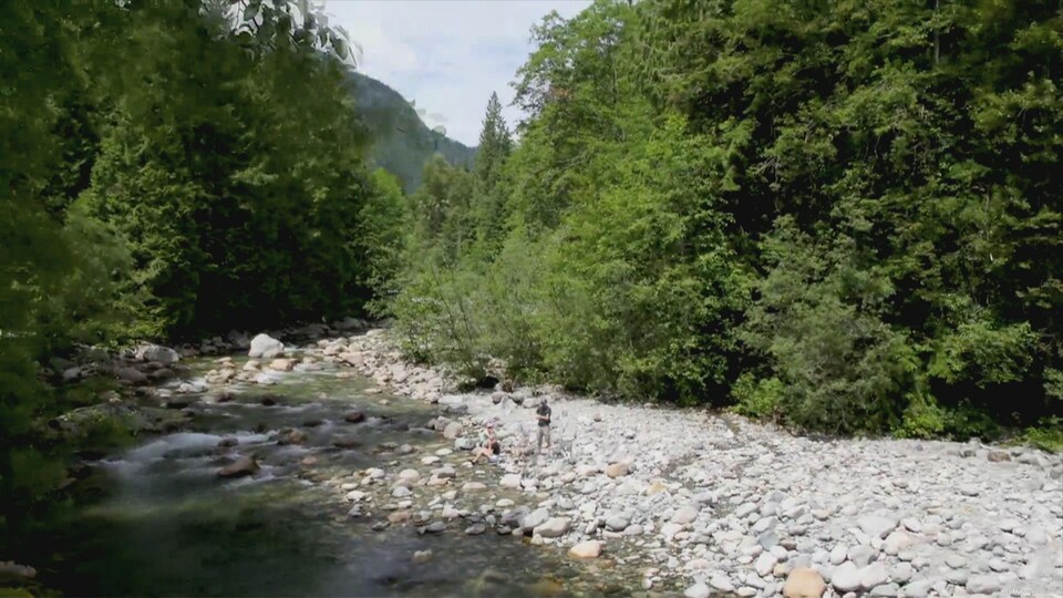 Deux personnes sont au bord d'une rivière peu profonde, entouré de rochers et bordés de grands conifères.