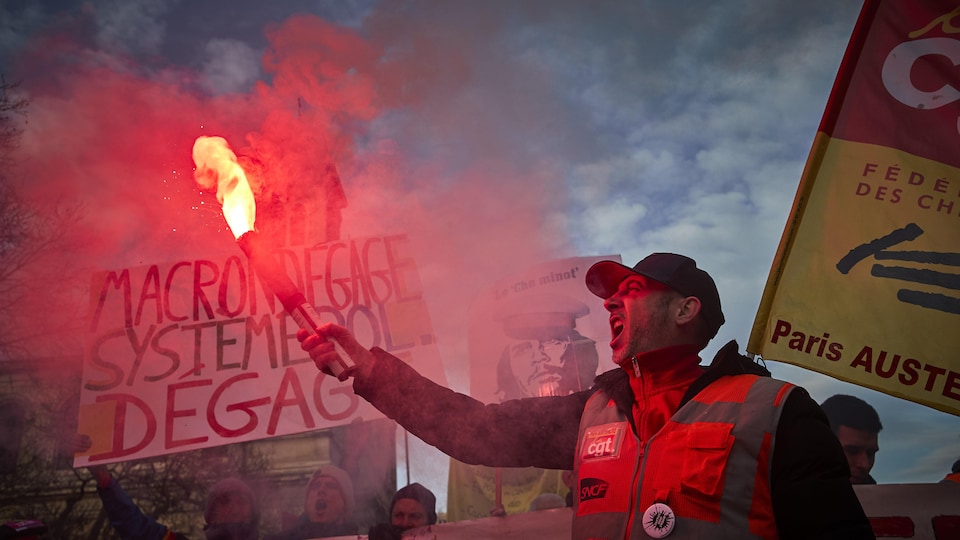 Un homme tient une fusée d'urgence allumée, des gens tiennent des affiches derrière lui.