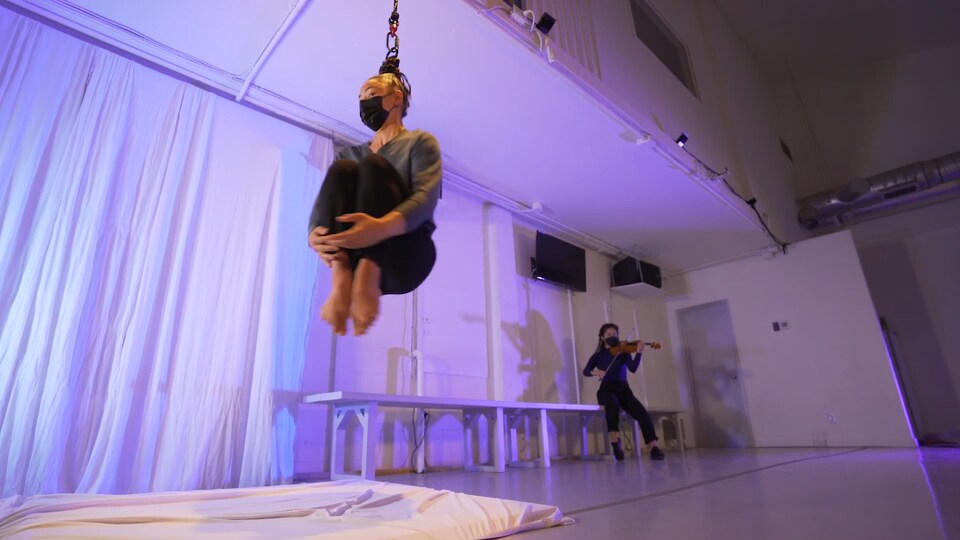 Une danseuse est attachée au plafond par les cheveux alors qu'une femme joue du violon en arrière-plan.