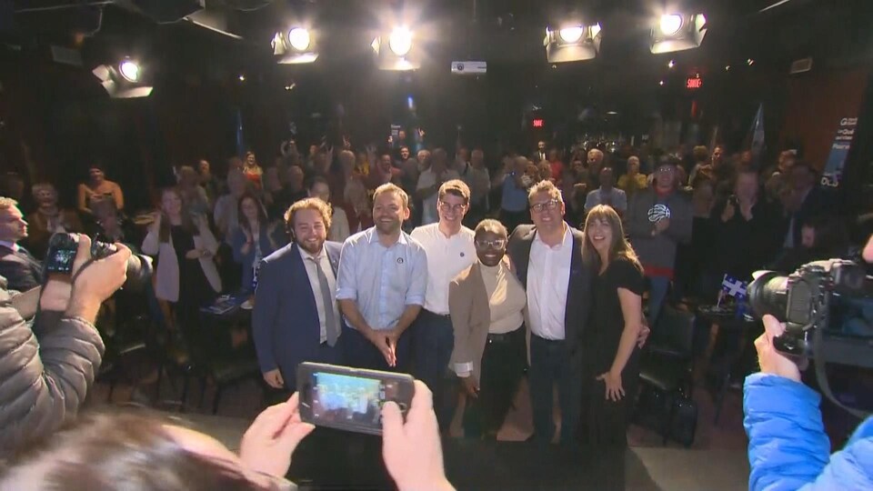 Une photo de groupe des candidats et du chef. Des dizaines de personnes à l'arrière-plan.