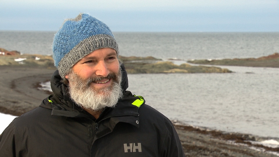Un homme barbu avec une tuque sourit pendant une entrevue sur une plage.