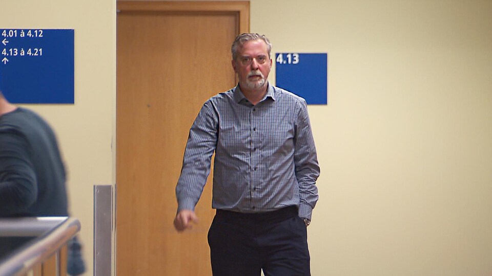 Monsieur Desgagnées est vêtu d'une chemise à carreaux de teinte bleue. Il marche dans le couloir.