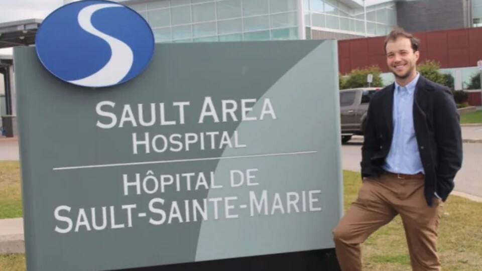 Lucas sourit à côté de l'enseigne de l'hôpital. Il a les mains dans ses poches.