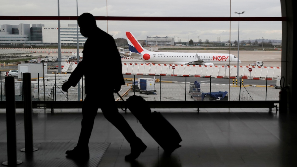 La silhouette d'un homme tirant une valise se profile devant une baie vitrée donnant sur le tarmac d'un aéroport.